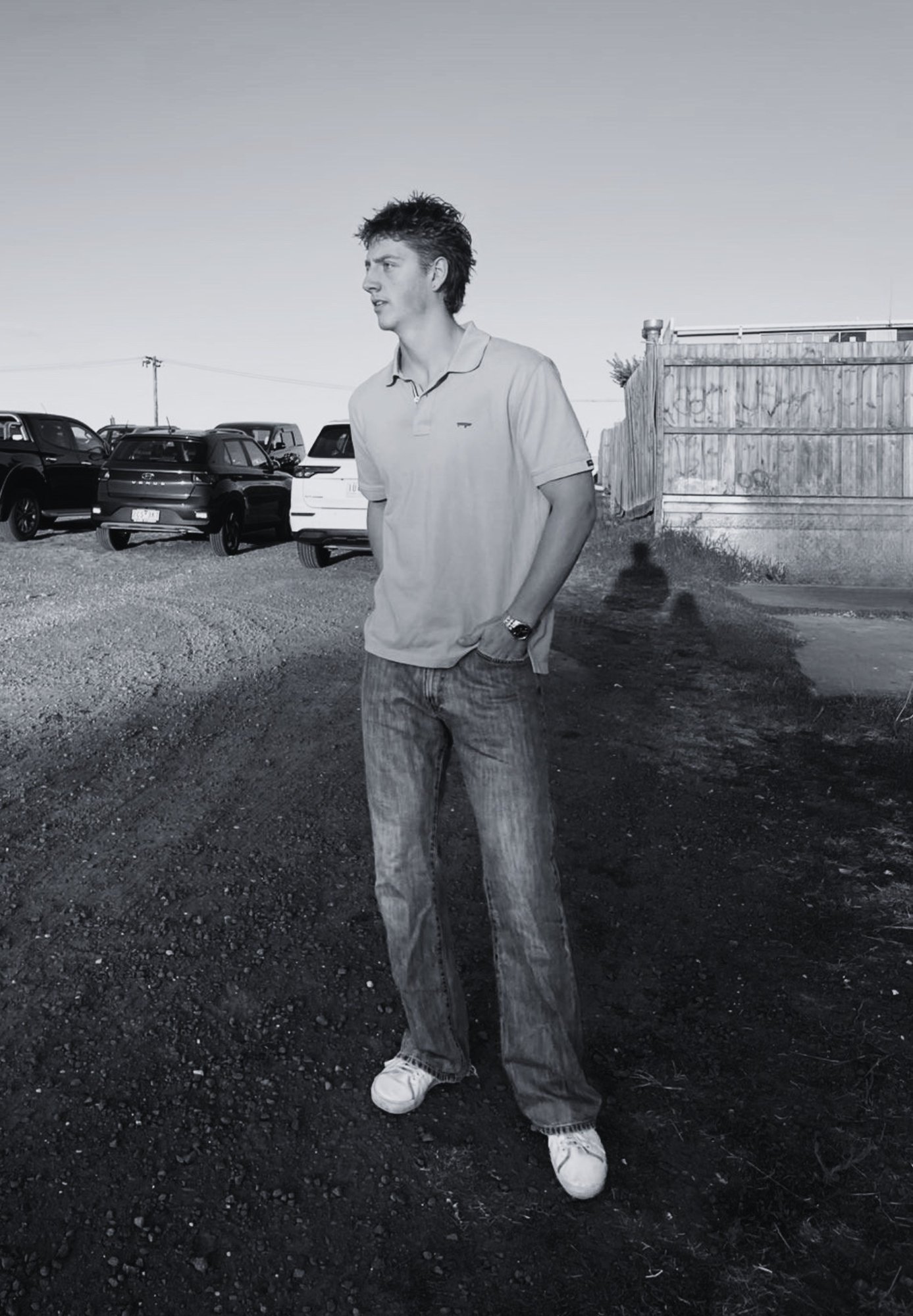 Young man standing in a junkyard wearing a polo shirt and jeans, looking to the side with parked cars and a metal gate in the background