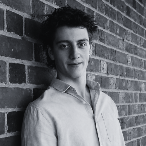 Black and white portrait of a young man with dark hair wearing a light button-up shirt, standing against a brick wall